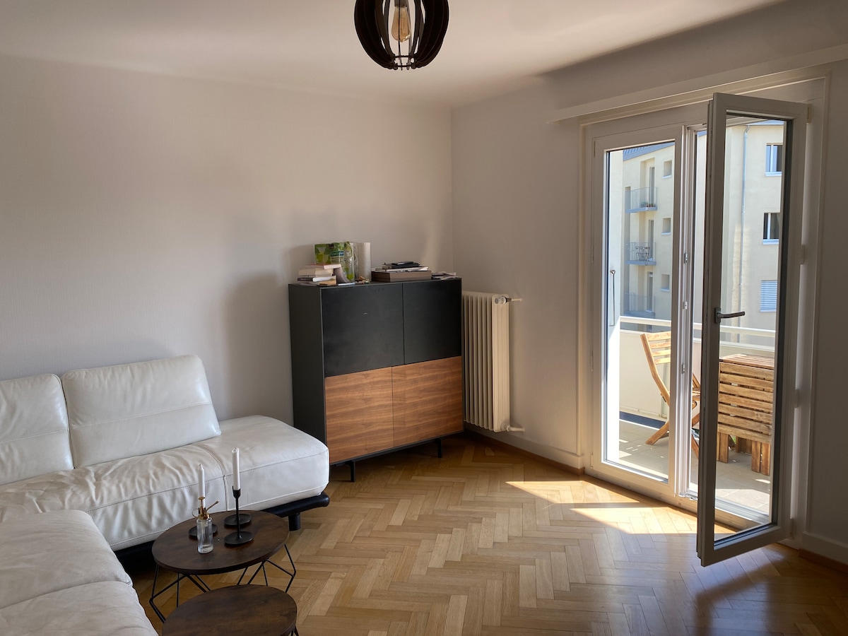 A bright living area features a white sectional sofa, complemented by a dark wooden cabinet. Light streams through the glass doors, which open to a balcony with outdoor seating. A small table and candlesticks add a touch of elegance to the space.