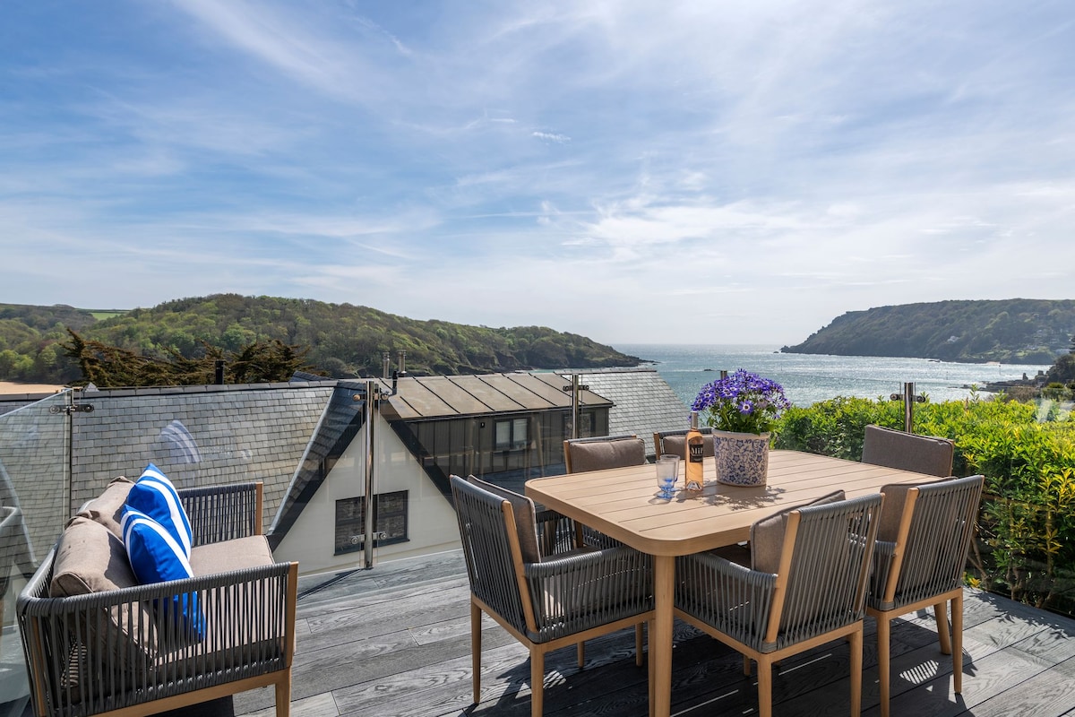 A private deck is shown, featuring an outdoor dining table surrounded by six cushioned chairs. A soft blue accent pillow rests on a nearby rattan chair. Expansive views of the estuary and coastline are visible in the background, complemented by a clear blue sky.