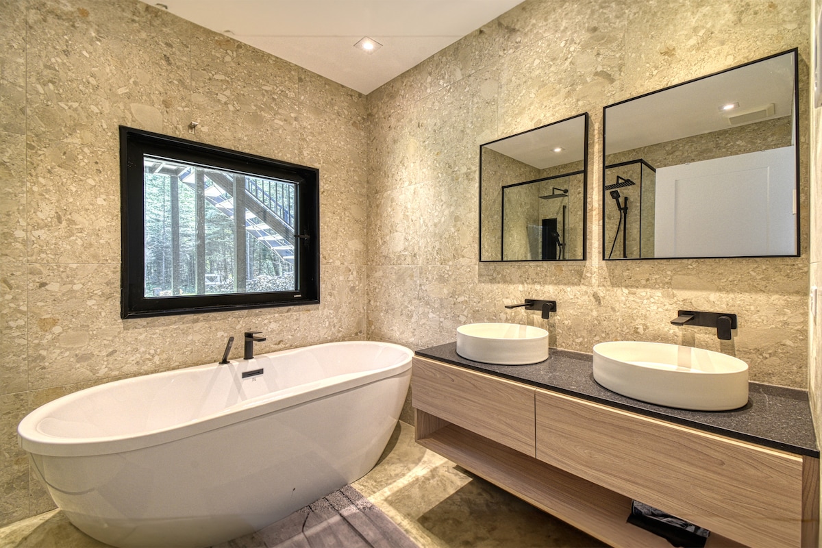 A modern bathroom features a freestanding oval tub beside a sleek double-sink vanity. Two circular basins are set on a dark countertop, complemented by minimalist black fixtures. Natural stone walls create a soothing backdrop, while a large window provides a view of the surrounding nature.