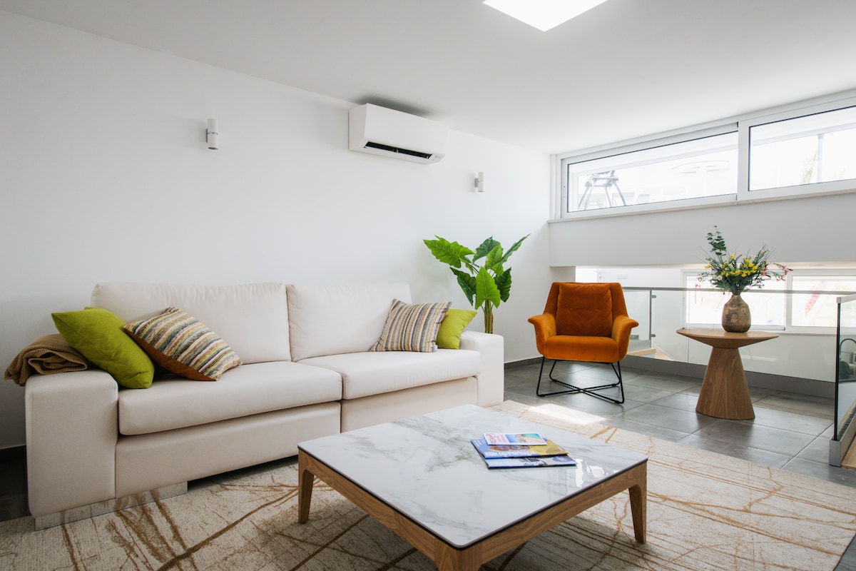A modern living area features a light-colored sofa adorned with decorative pillows and a cozy orange armchair. A wooden coffee table sits on a neutral rug, while a potted plant adds a touch of greenery. Natural light floods the space through large windows.