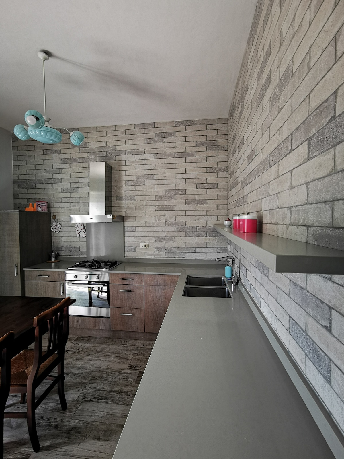 A modern kitchen features brick-patterned walls and a sleek gray countertop. Stainless steel appliances, including a gas stove and a hood, are integrated into the design. A wooden dining table is present, along with dark wooden chairs, creating a functional cooking and dining space.