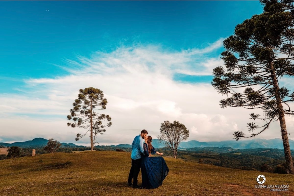A couple is embraced on a grassy hillside, surrounded by rolling mountains beneath a vast blue sky. Tree silhouettes frame the scene, enhancing the natural beauty. The sunlight casts soft shadows, creating a peaceful setting that invites tranquility and connection with nature.