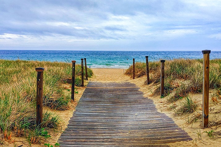 A wooden walkway leads from a sandy path through tall grass to a serene beach. The ocean appears calm under a cloudy sky, with gentle waves lapping at the shore in the distance.