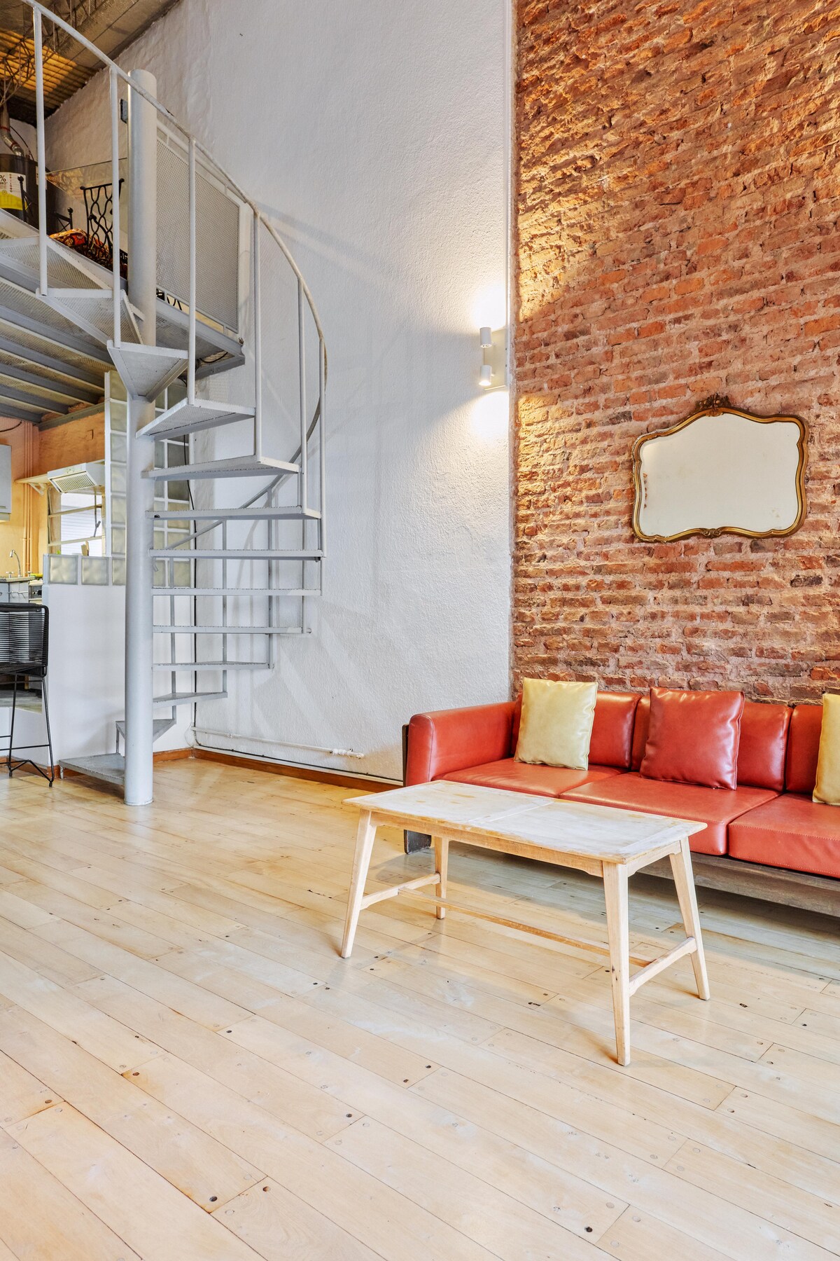 A spacious living area features a vibrant red sofa adorned with cushions. A simple wooden coffee table occupies the space, while a spiral staircase leads to the upper level. Exposed brick walls and polished wooden floors contribute to the modern loft's charm.