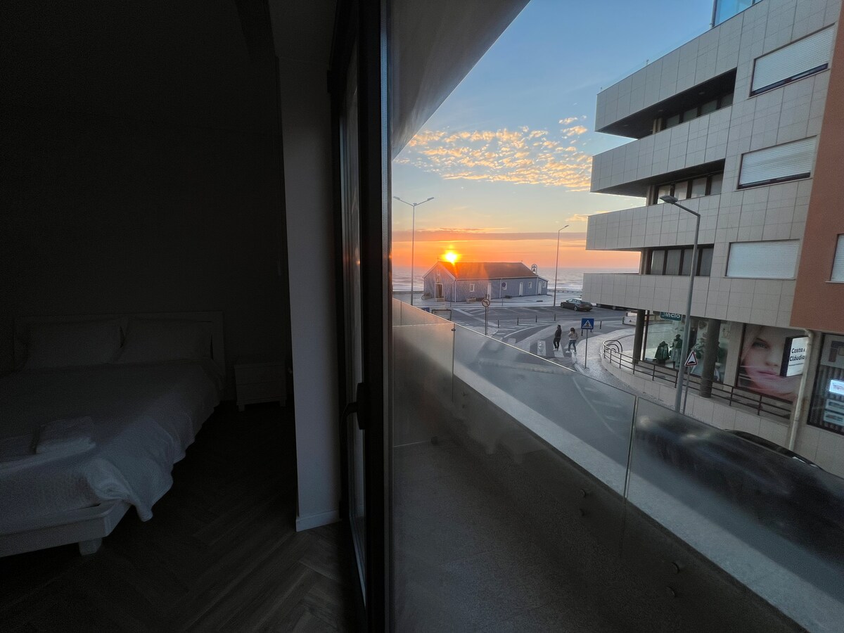 A serene view of the ocean at sunset is visible through large glass doors. The warm hues of the sky contrast with the modern architecture nearby. A spacious bed can be seen from the interior, creating a peaceful atmosphere.