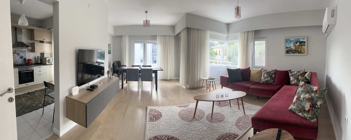 A spacious living area is featured, offering a cozy sectional sofa adorned with patterned cushions. A wooden coffee table sits centrally, and a dining table is positioned nearby. Large windows allow natural light to fill the space, complemented by soft curtains.