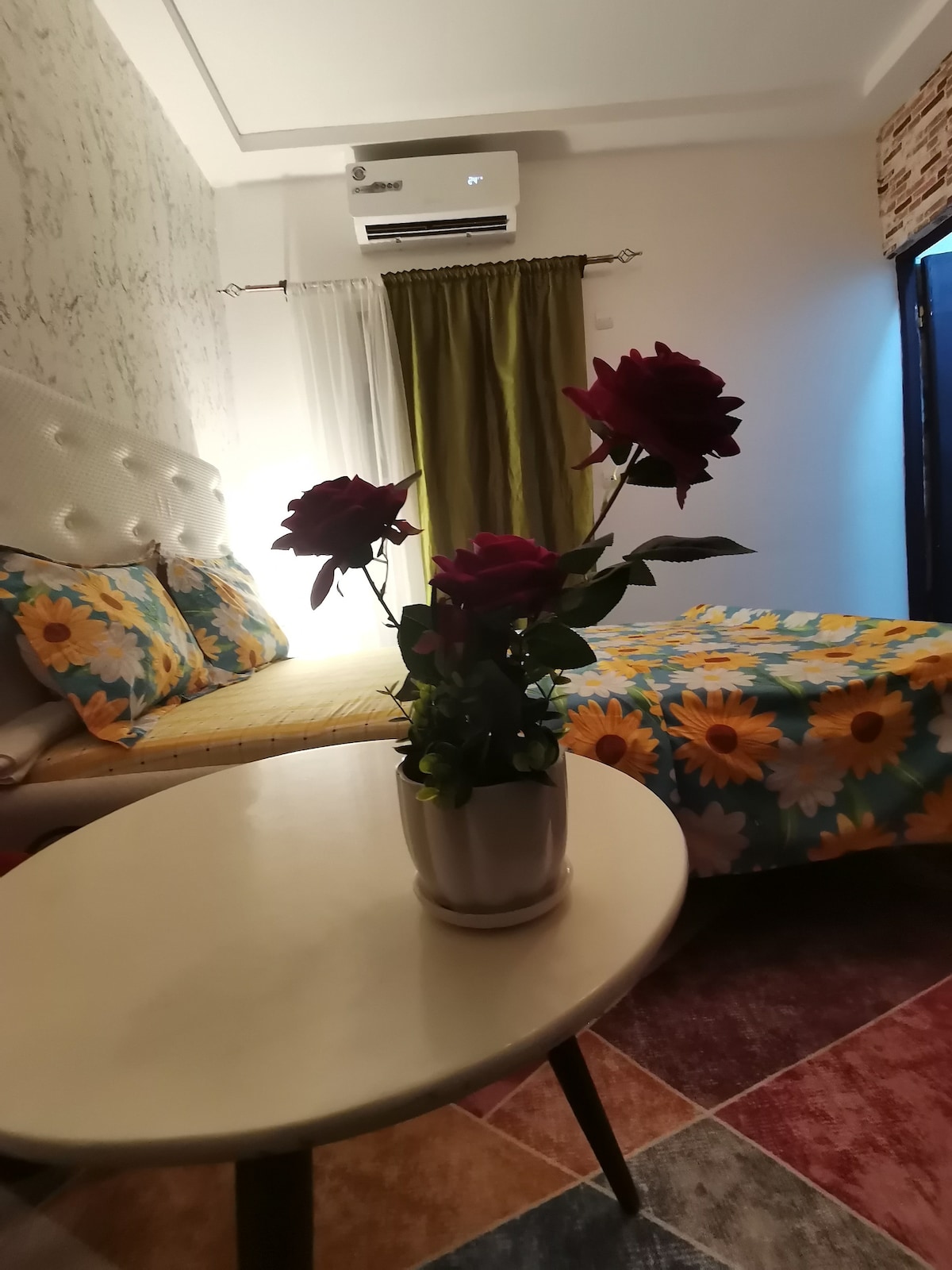 A cozy bedroom features a spacious bed adorned with a sunflower-patterned blanket. Decorative floral pillows complement the bed. A small table with a vase of vibrant roses is positioned in front of the bed. Soft curtains frame a window that provides natural light.