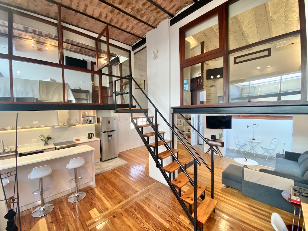 An open-plan loft is featured, showcasing a modern kitchen with sleek appliances. Spacious seating areas are visible, alongside wooden floors and an elegant staircase leading to an upper level. Large windows allow natural light to fill the space, enhancing the overall atmosphere.