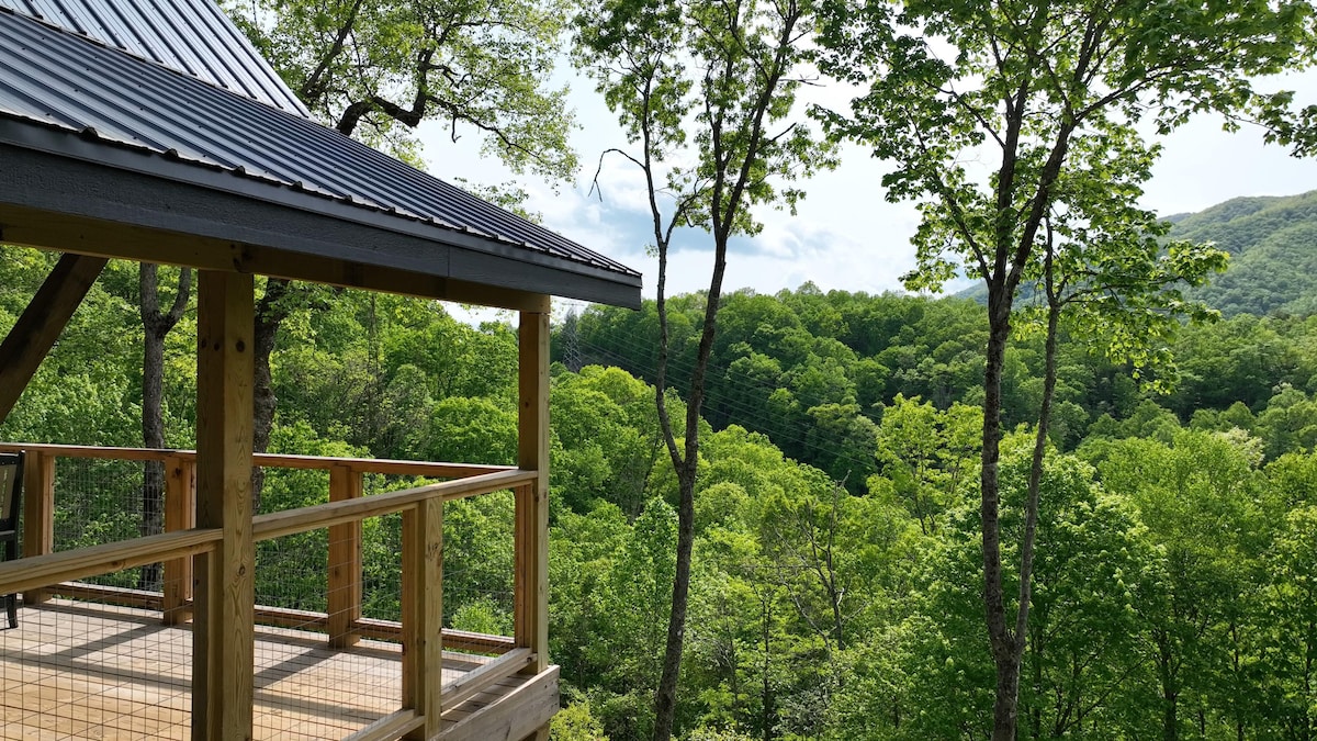 A spacious deck with wooden railings is visible, providing unobstructed views of the lush green landscape. Tall trees frame the scene, while distant mountains offer a serene backdrop. Natural light illuminates the space, highlighting the cabin's connection to the surrounding nature.