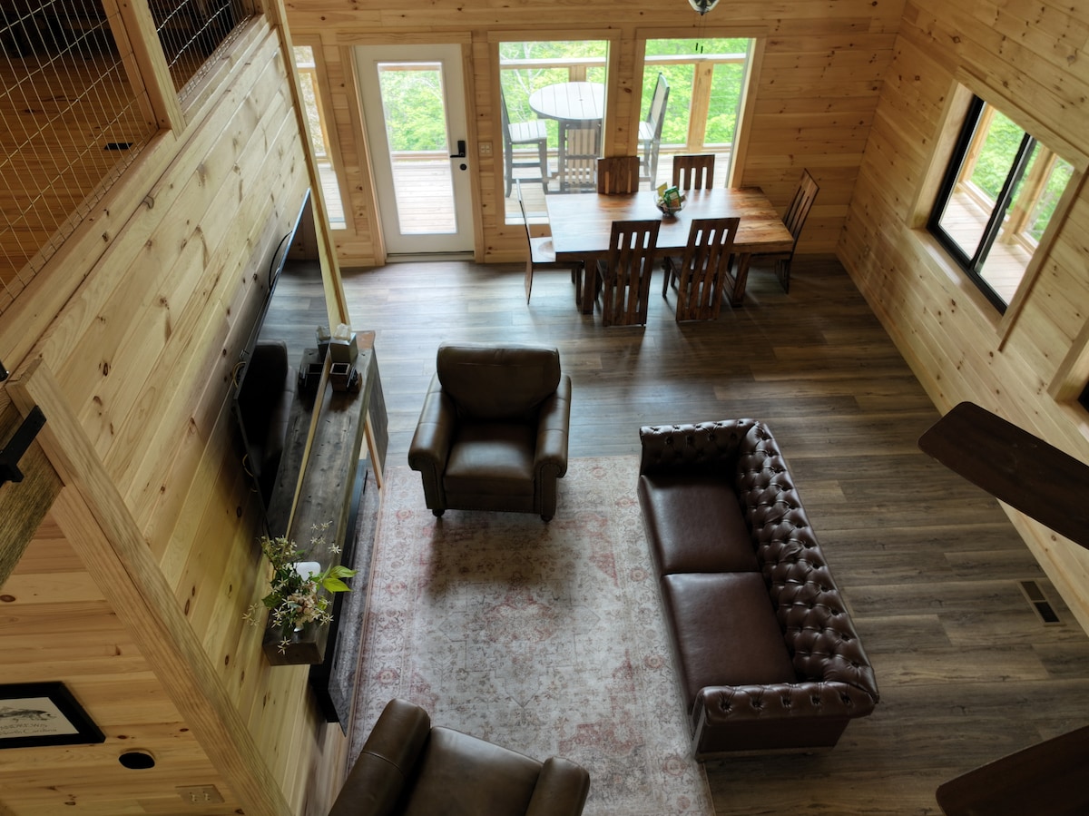 An aerial view of the open-concept living area reveals rustic wooden walls and flooring. A cozy seating arrangement includes two leather chairs and a plush sofa. A dining table is seen in the background, surrounded by wooden chairs, with doors opening to a deck offering natural light.