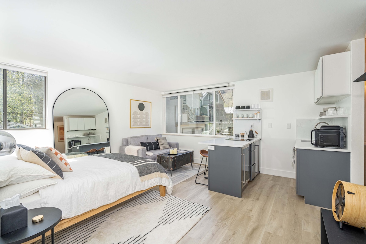 A bright, modern studio space features a queen bed with neutral linens and decorative pillows. A cozy seating area with a sofa is present beside a kitchenette equipped with essential appliances, including a microwave and coffee maker. A large window allows natural light and outdoor views.