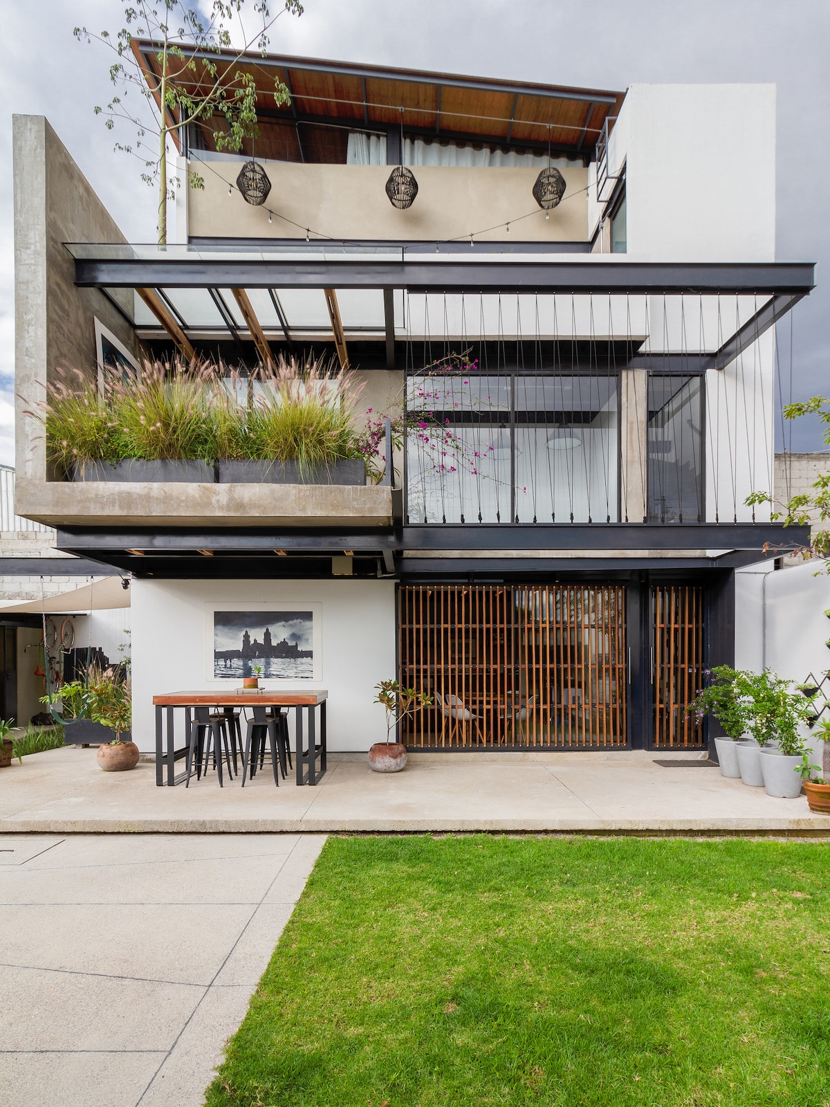 The building showcases a modern industrial design with multiple levels and large windows. A private terrace is visible on the upper floor, adorned with greenery. A spacious outdoor area includes a dining table and chairs, bordered by a well-maintained lawn.