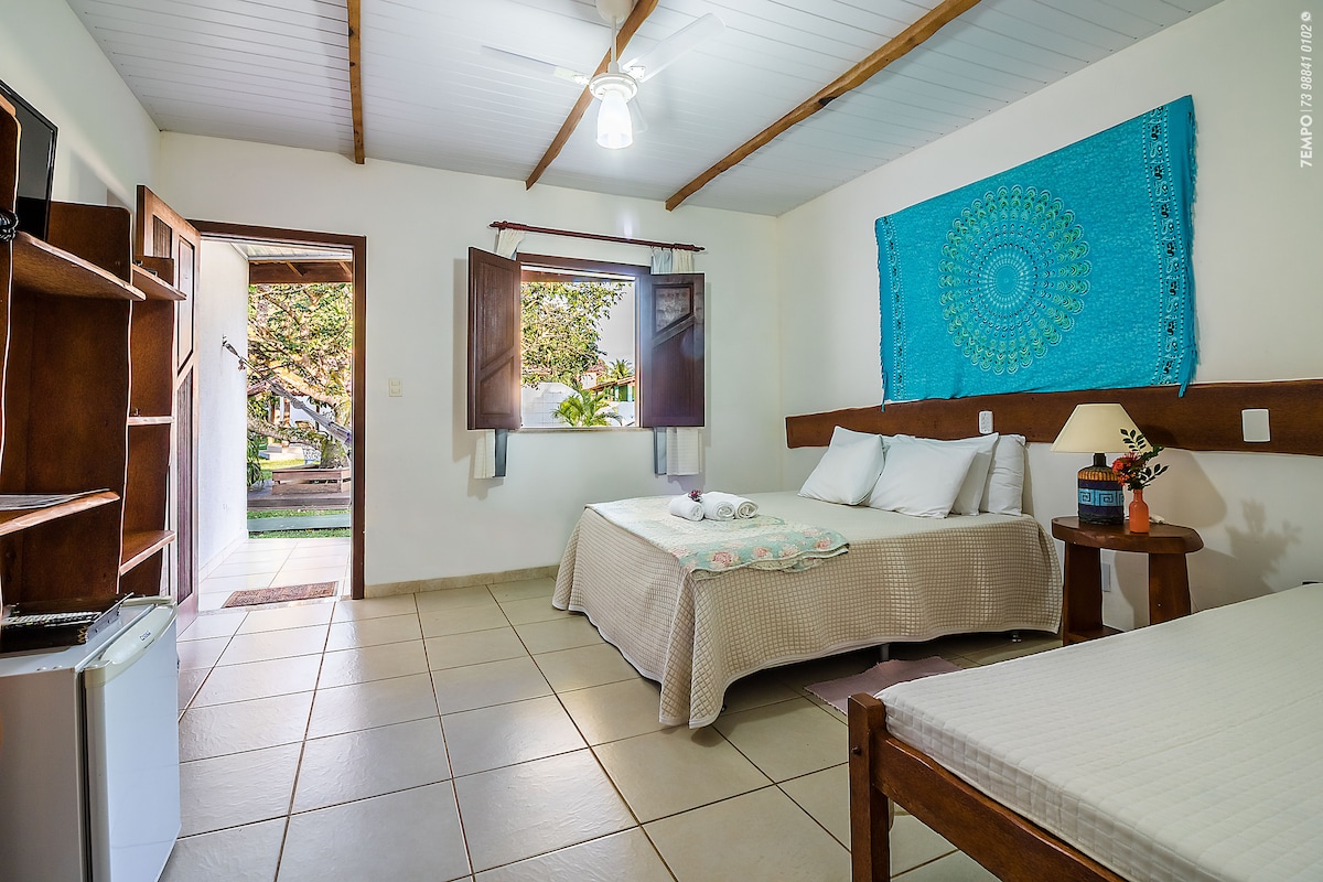 A bright and airy guest room features two beds, one larger and one smaller, with neutral bedding. A colorful wall hanging provides a pop of color. Open windows allow natural light and outdoor views. A small refrigerator and shelving unit are included in the space.