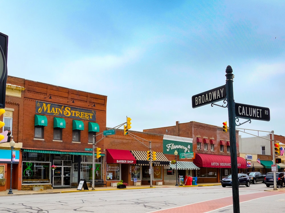 A charming street view is displayed, featuring brick buildings with awnings in various colors. Business signs are visible, indicating shops and services, while traffic signals and street signs denote the intersection of Broadway and Calumet Road. A wide sidewalk invites pedestrian activity.