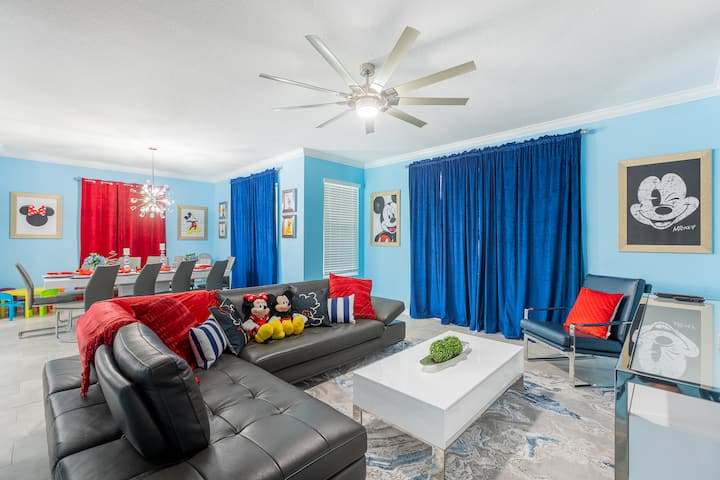 ก้าวเข้าสู่ห้องนั่งเล่นและพื้นที่รับประทานอาหารแบบเปิดโล่งที่สดใสนี้! ผนังสีฟ้าสดใสและผ้าม่านสีแดงเข้มสร้างบรรยากาศสไตล์ดิสนีย์ที่ดูสนุกสนาน จับคู่กับที่นั่งสีดำโฉบเฉี่ยว โต๊ะกาแฟสีขาวทันสมัย งานศิลปะมิกกี้ และพื้นที่รับประทานอาหารสไตล์ครอบครัวที่น่าดึงดูด  