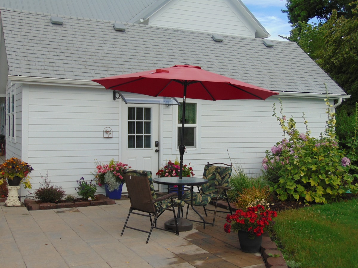 An outdoor patio space is shown, featuring a round table with two chairs under a red umbrella. Colorful flower pots line the edges, with vibrant blooms and greenery enhancing the scene. The white building with a gray roof stands in the background.