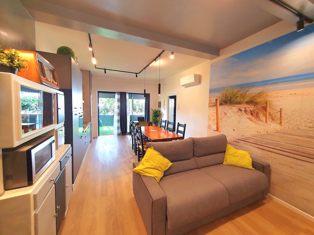 The living area is designed with an industrial-chic style, featuring a gray sofa and a dining table that accommodates eight. A large wall mural depicts a beach scene, while glass doors lead to an outdoor space. Natural light enhances the bright wooden floors.