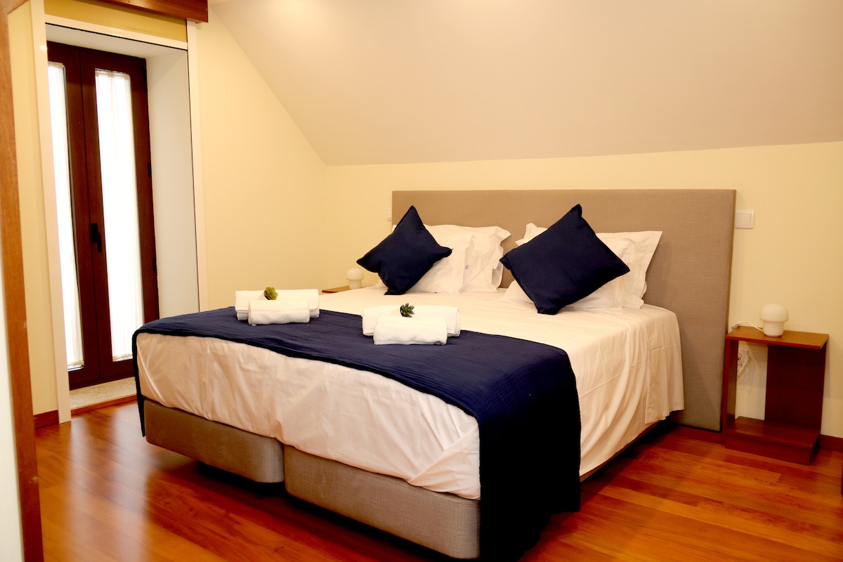 A spacious sleeping area features a comfortable bed dressed in neutral linens, accented by dark blue pillows. Fresh towels are neatly arranged on the bed. A wooden nightstand is placed beside the bed, and natural light enters through a window with wooden shutters.