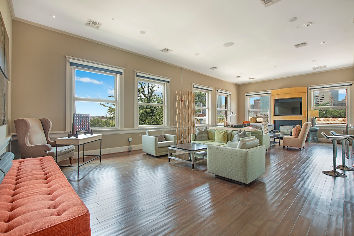 A spacious common area is equipped with a variety of seating options, including a coral-colored sofa and light green armchairs. Large windows allow natural light to fill the space, while a sleek coffee table sits at the center. The room features wood flooring and modern decor.