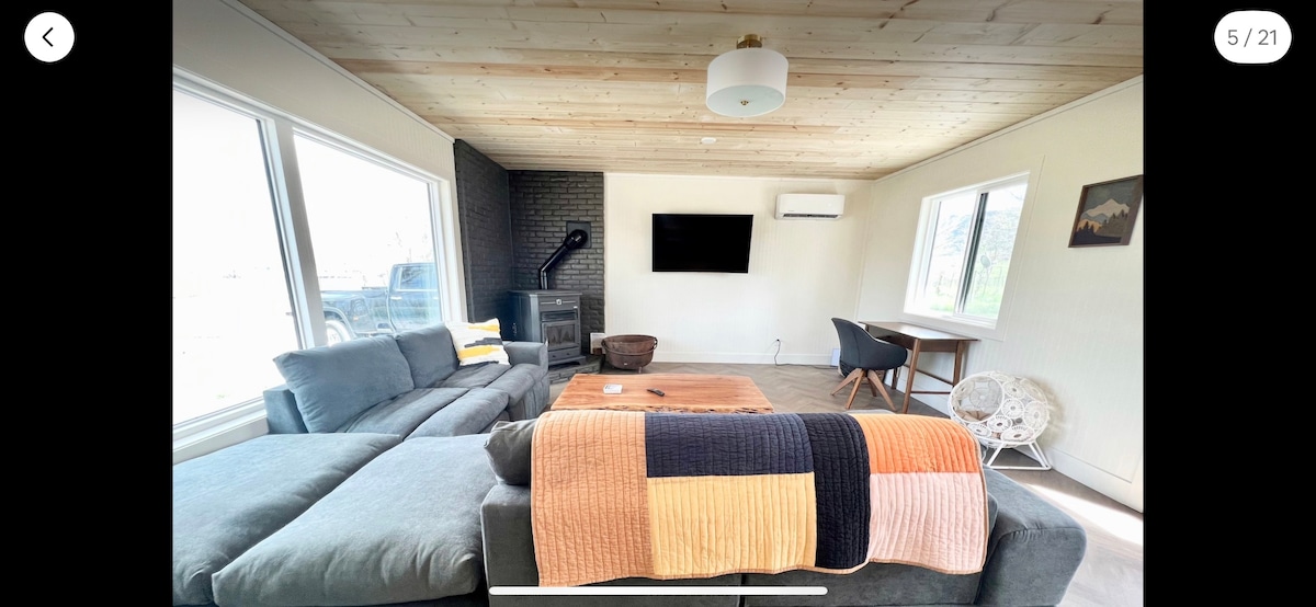 A spacious living area features large windows allowing natural light to flood in. A sectional sofa is paired with a contrasting throw blanket. A flat-screen TV is mounted on the wall, and a small dining table is positioned nearby, complemented by a modern chair.