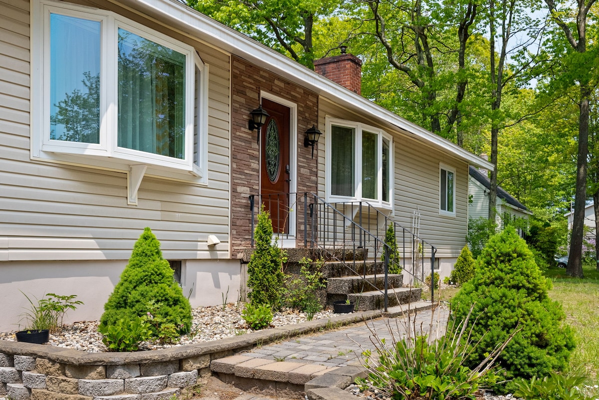 The front exterior of the home is depicted, showcasing a welcoming entrance with stone steps leading to a door framed by decorative lighting. Symmetrical greenery, including carefully shaped shrubs, adds a natural touch, while large windows provide glimpses of the interior.