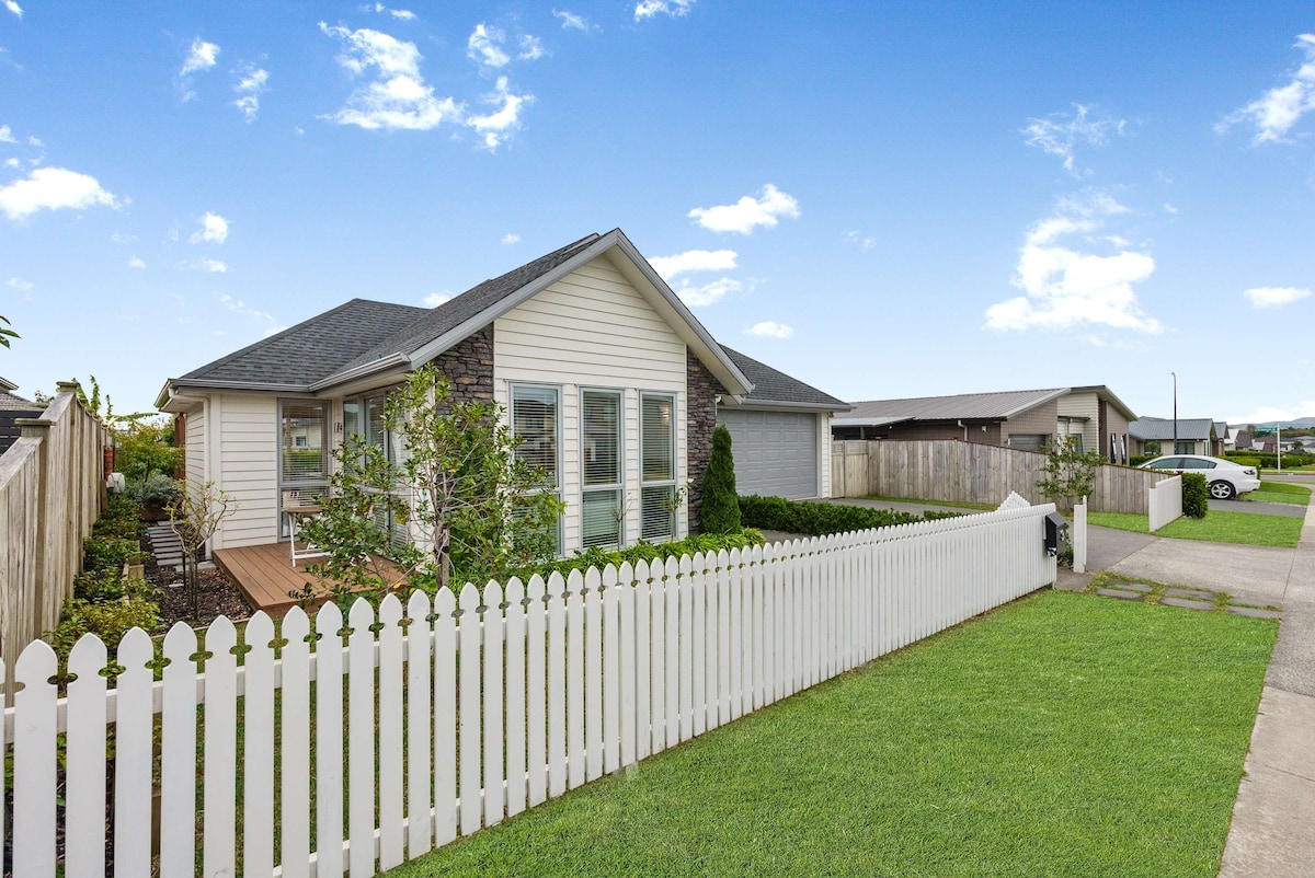 A charming house is framed by a white picket fence, with a well-kept lawn and landscaped greenery. The exterior features a mix of stone and wooden elements, complemented by large windows. A driveway leads to a nearby vehicle parked on a concrete surface.