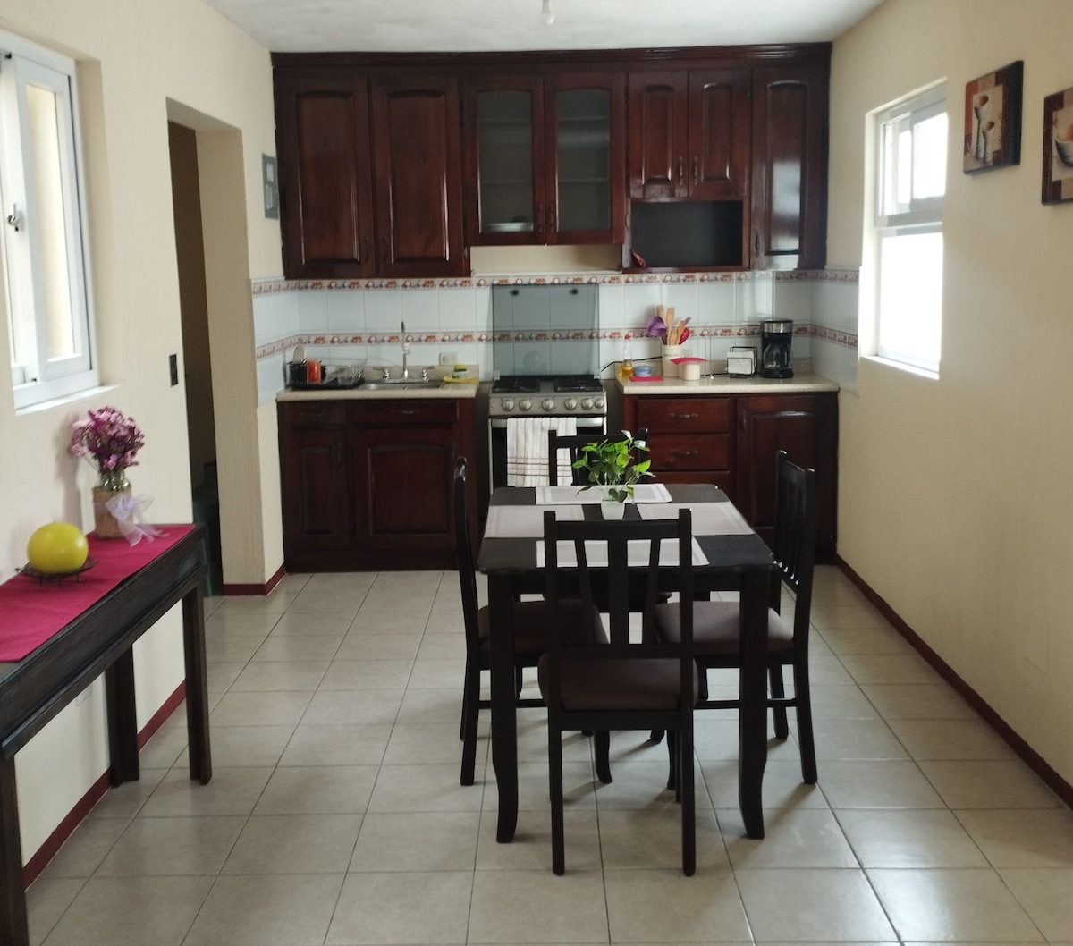 A spacious kitchen area is furnished with dark wooden cabinetry and a countertop. A dining table with four chairs is positioned in the center, complemented by a floral centerpiece. Natural light enters through two windows, enhancing the warm tones of the room.