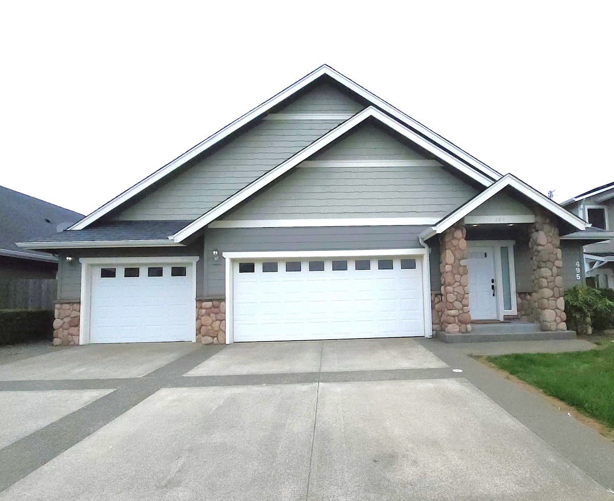 A contemporary house is showcased with a large, gray exterior and stylish stone accents. The front features a covered entryway and a three-car garage with white doors. A well-maintained lawn is visible, bordered by a neatly paved driveway.