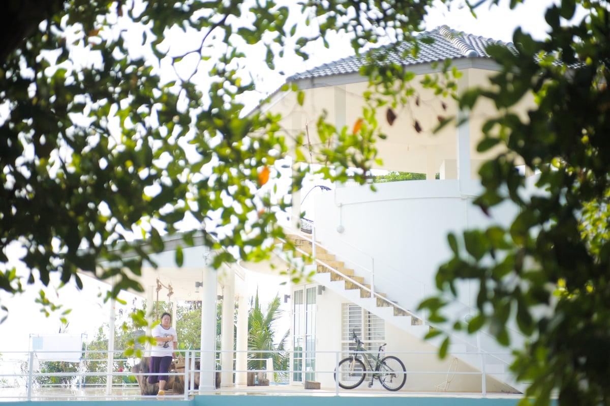 A view of the holiday home is framed by lush greenery, showcasing a spacious upper patio supported by a sturdy white railing. A bicycle leans against the home, and a figure stands on the patio, surrounded by palm trees and the serene landscape of the bay.