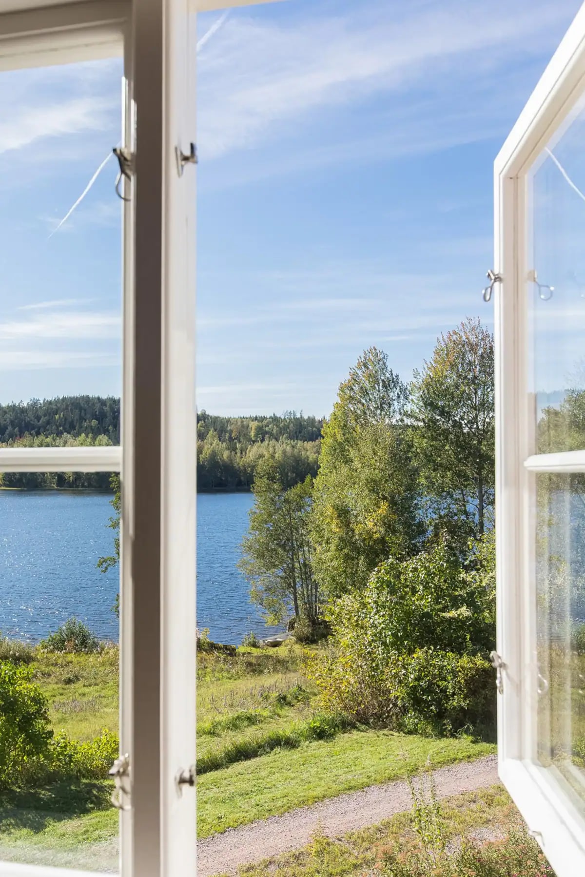 The view from an open window frames a serene lake surrounded by lush greenery and trees. The soft blue water reflects the clear sky, while the landscape features a gentle path leading through the grassy foreground.