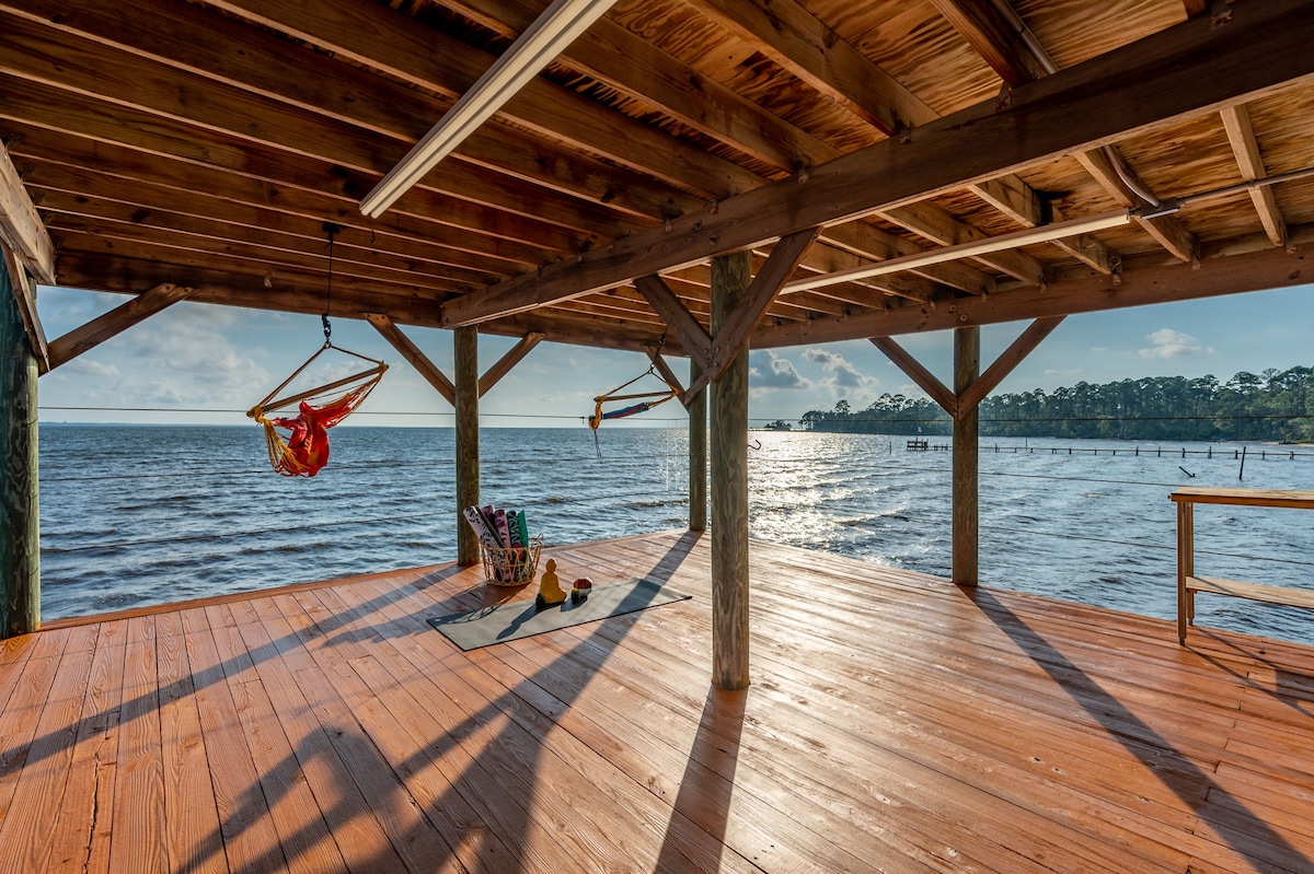 A spacious two-story dock extends over the water, featuring an inviting wooden deck with ample room for relaxation. A hammock is suspended nearby, alongside paddleboards and yoga mats organized in a neat arrangement, while the shimmering water reflects the warm light of the setting sun.