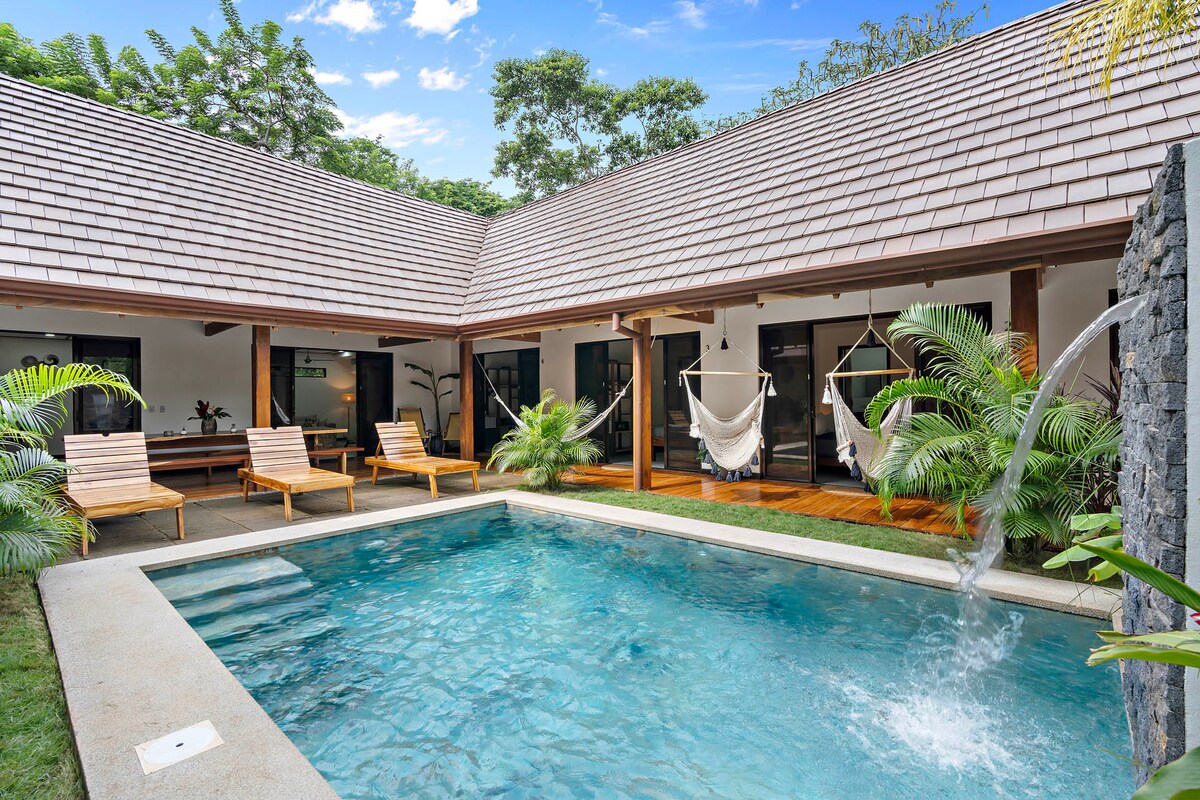 A tranquil outdoor pool is surrounded by a U-shaped layout of rooms, with wooden lounge chairs placed beside it. Lush greenery is visible, along with two hammocks hanging from the structure. The scene is brightened by natural light filtering through the surrounding foliage.
