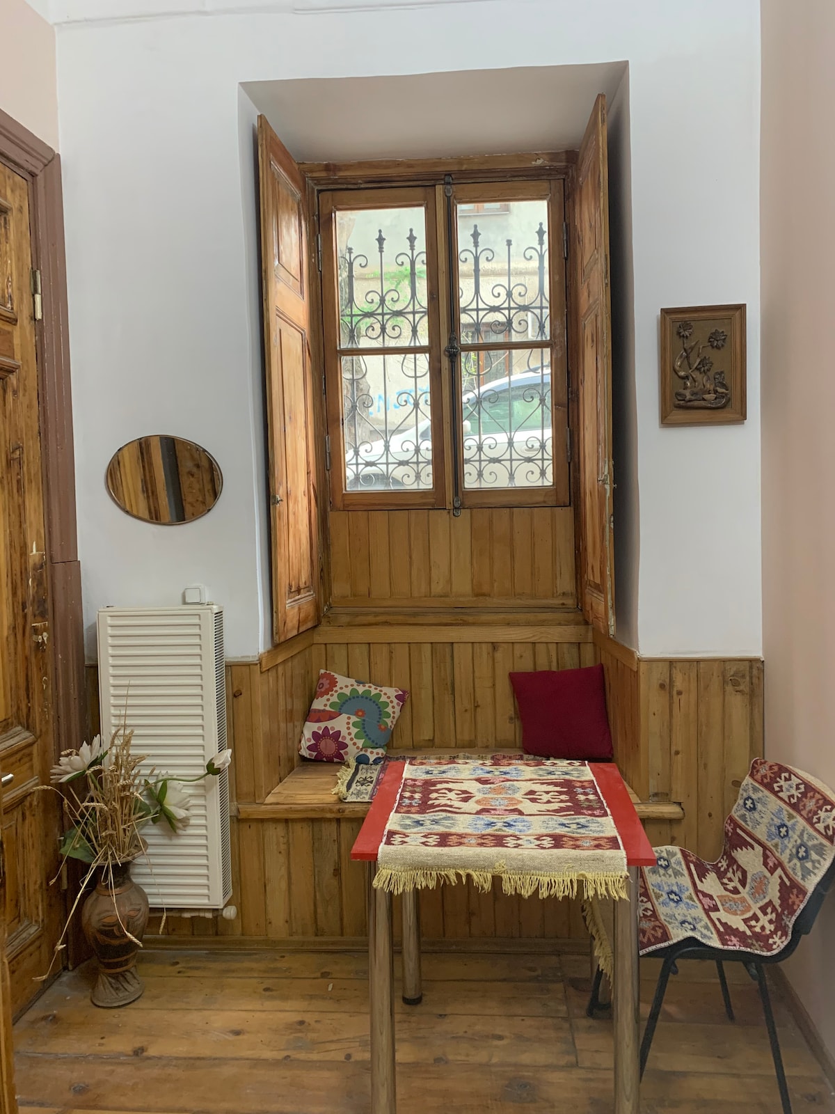 A cozy nook features a wooden bench flanked by decorative cushions, with a small table covered in a colorful tablecloth. Light enters through a window adorned with intricate ironwork, while wooden paneling adds warmth to the space.
