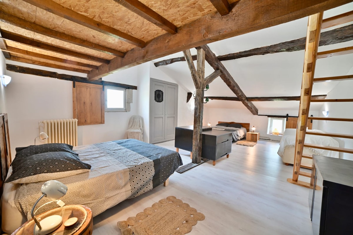 The spacious bedroom features exposed wooden beams and natural light streaming through a small window. Two beds with neutral bedding are positioned beside each other, along with a small bedside table. A cozy area rug lies beneath, enhancing the room's charm.