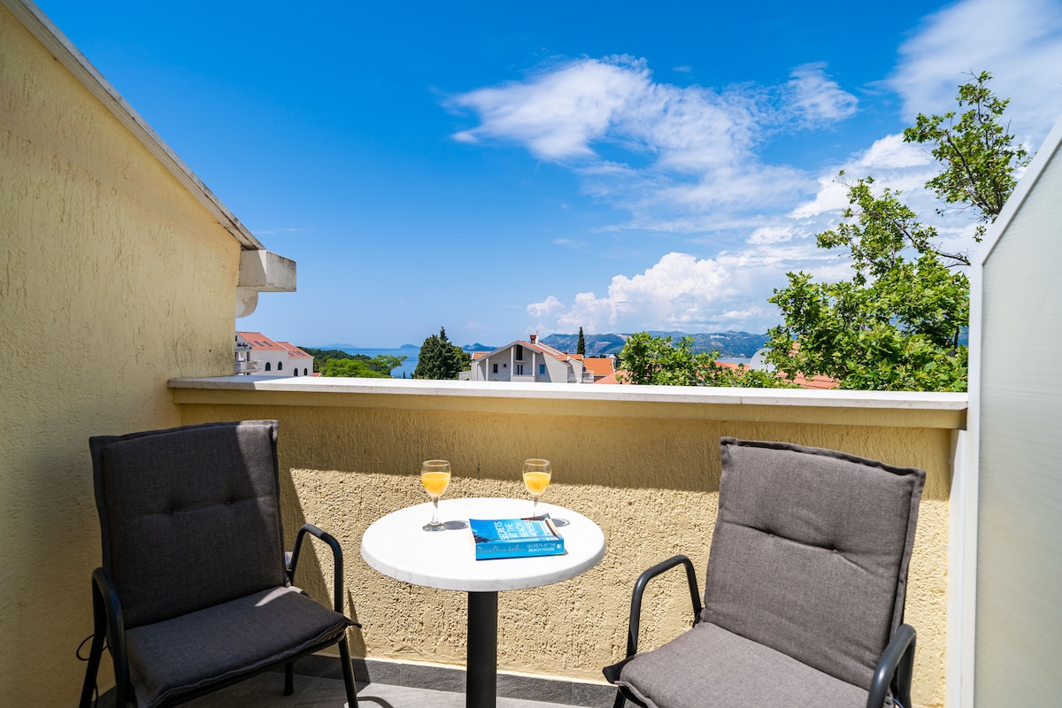 A private balcony features two cushioned chairs and a small round table, set with two drinks. The view includes distant landscapes and nearby buildings under a clear blue sky, providing a serene outdoor space for relaxation.