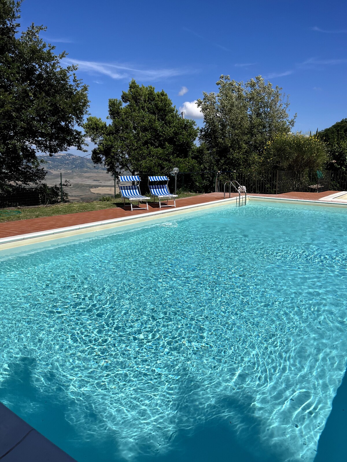 A serene communal pool showcases clear blue water, reflecting sunlight and surrounded by lush greenery. Two blue and white striped lounge chairs are positioned nearby, inviting relaxation. The backdrop reveals rolling hills, enhancing the tranquil outdoor setting.