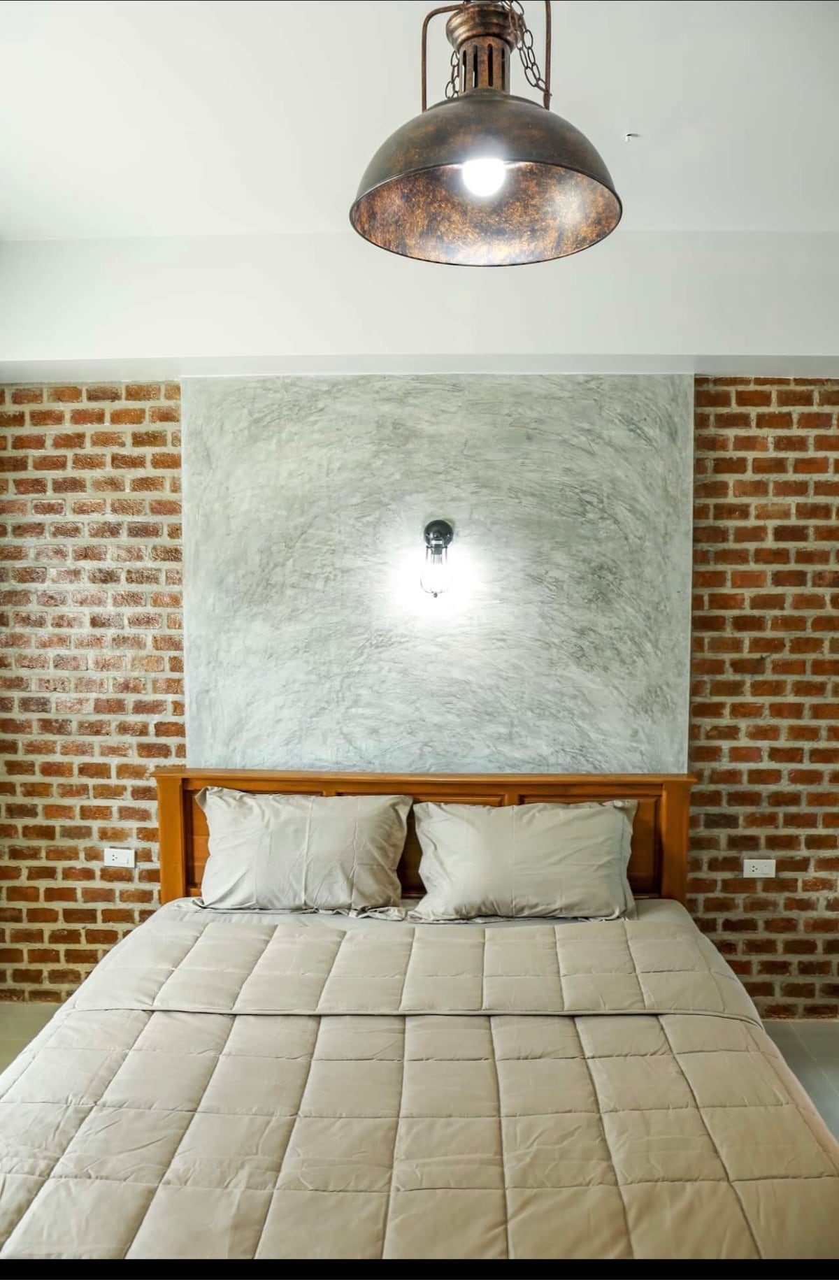 A cozy sleeping area features a wooden bed with soft, neutral bedding and two matching pillows. A textured gray wall is complemented by a rustic brick accent wall. A simple pendant light hangs overhead, providing soft illumination.