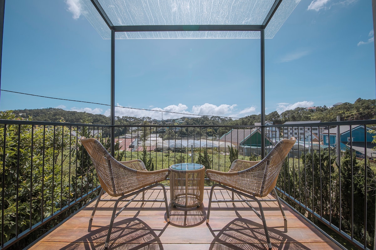 A spacious balcony is furnished with two wicker chairs and a glass table, offering a scenic view of the lush surrounding valley and gardens. Clear blue skies create a bright atmosphere, enhancing the outdoor experience.