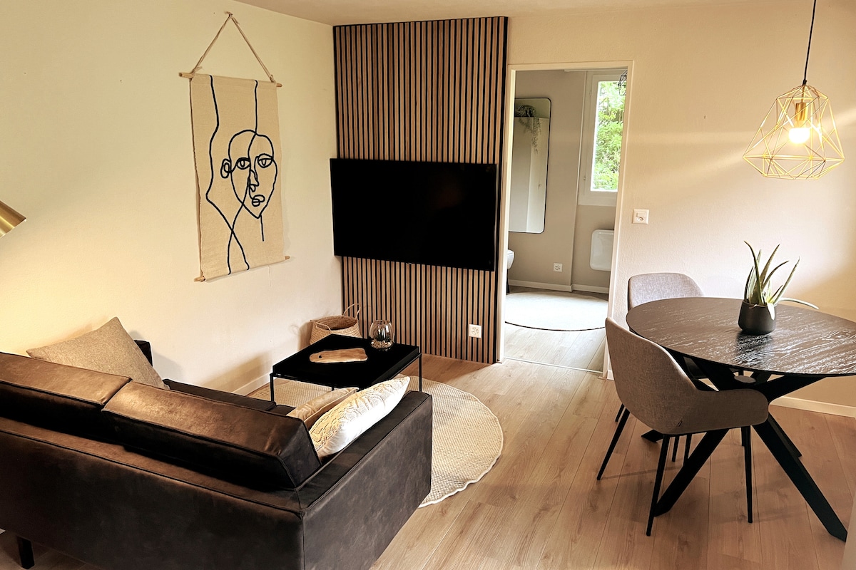 A cozy living area is displayed with a dark sofa facing a flat-screen television mounted on the wall. A small round dining table with gray chairs occupies one corner, while a decorative wall hanging adds a touch of character to the space.