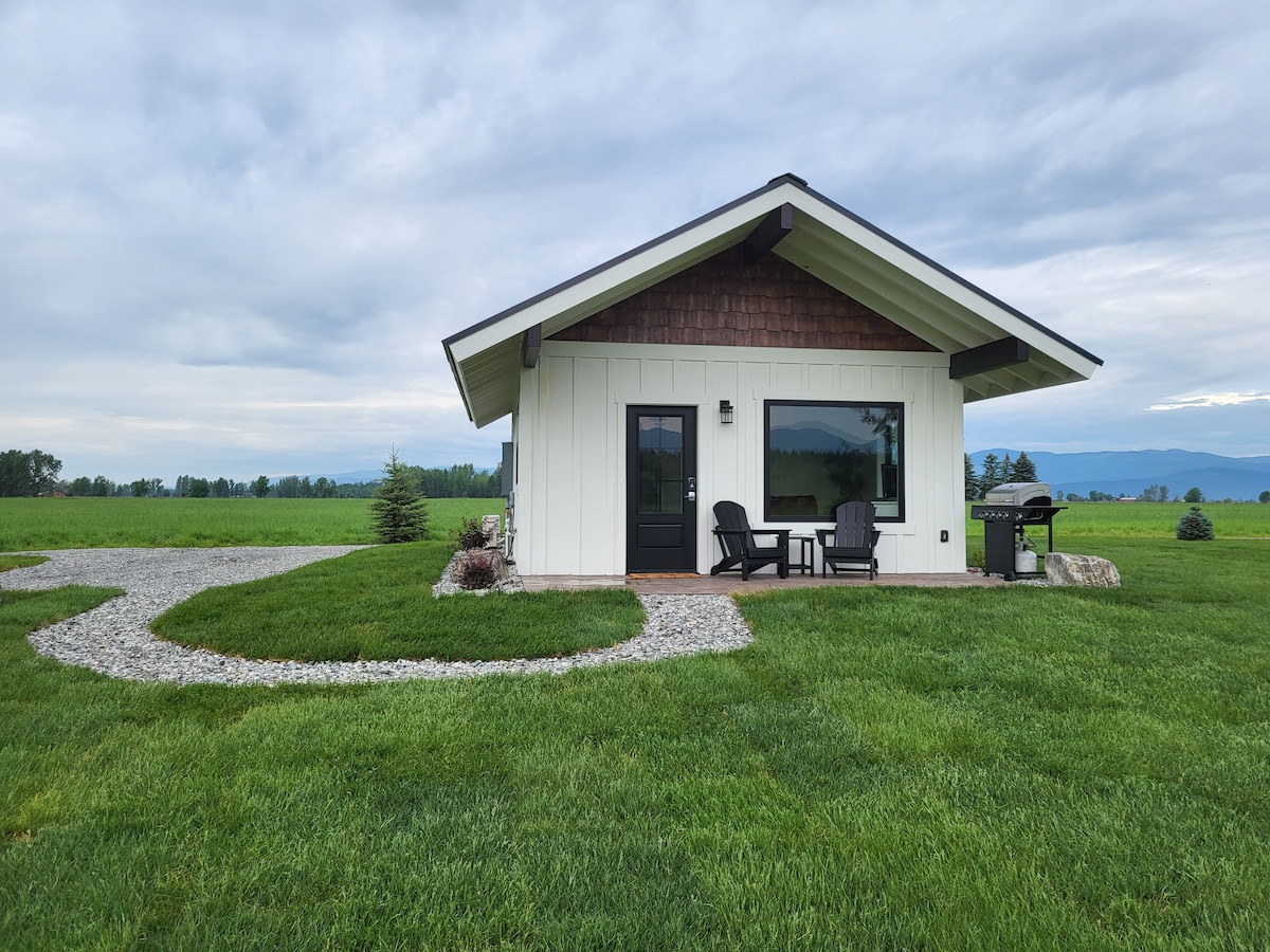 A modern cabin is set amidst expansive green grass, featuring a sloped roof and a combination of wooden and white exterior. Adirondack chairs are arranged on the porch, and a propane grill is positioned nearby. A stone pathway leads toward the cabin from the gravel driveway.