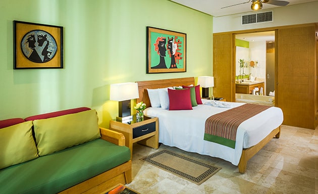 A spacious bedroom features a king bed with decorative pillows and a colorful throw blanket. A cozy sofa sits against the wall, complemented by art pieces. Soft lighting illuminates the room, while a doorway leads to a private bathroom area.