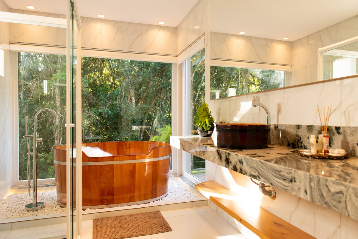 A sleek bathroom features a wooden soaking tub surrounded by large windows that offer views of the surrounding nature. The marble countertop provides space for toiletries and décor, while natural light fills the area, enhancing the serene atmosphere.