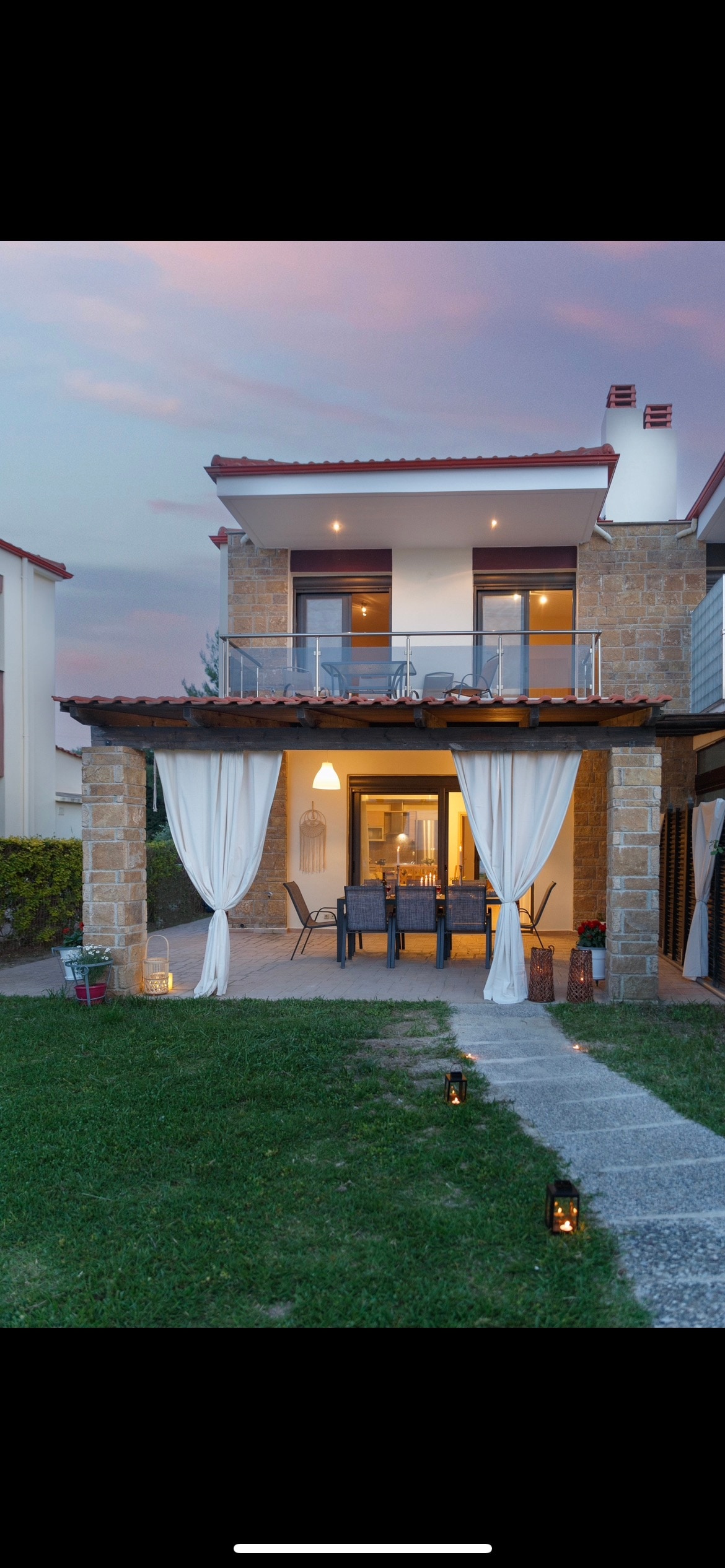 An inviting two-story residence features a balcony overlooking a grassy area. The ground level includes a covered outdoor seating area with a dining table and chairs, surrounded by soft curtains. Decorative lanterns line the pathway, leading to the entrance of the home.
