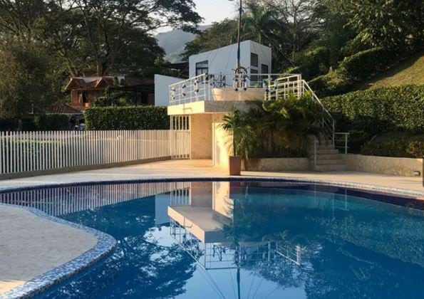 A tranquil pool area is presented, featuring clear blue water that reflects surrounding greenery. A spacious deck, complemented by a decorative plant in a pot, is visible along with a white structure overlooking the pool, enhancing the serene atmosphere of the outdoor space.