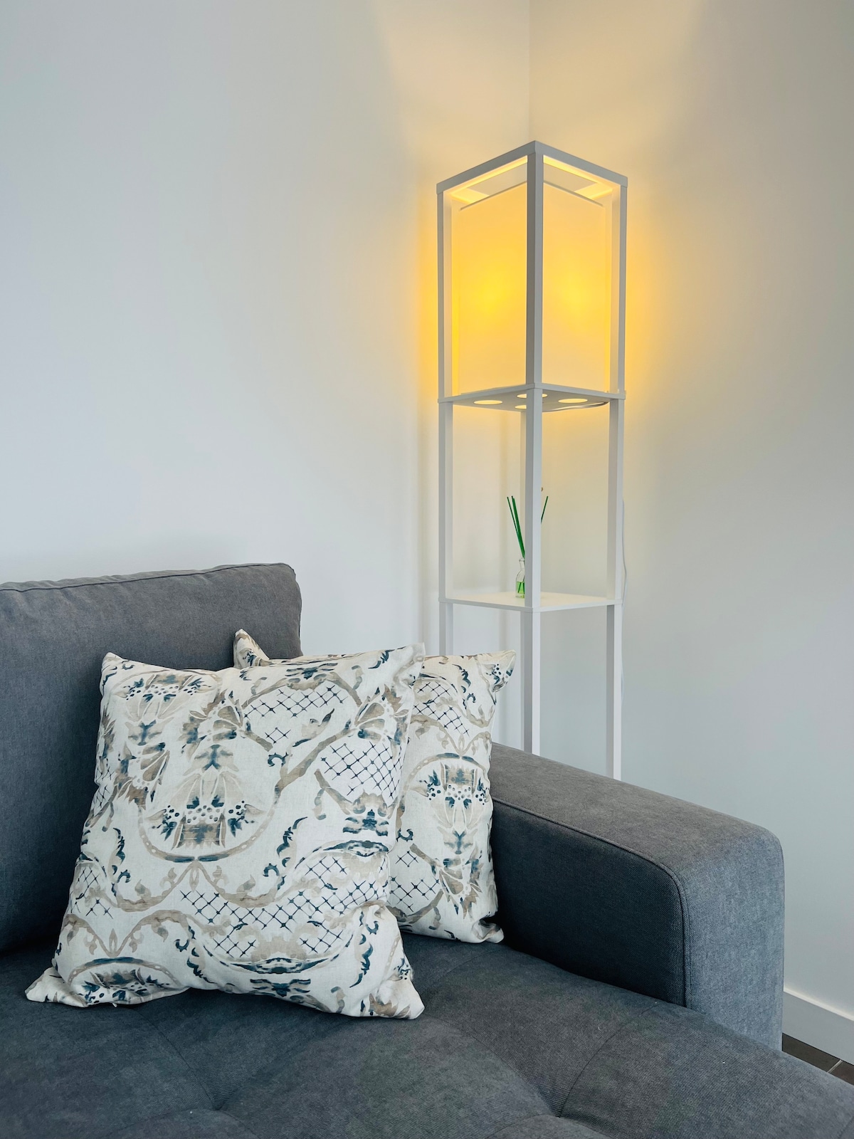 A corner of a living area features a gray upholstered sofa adorned with decorative throw pillows in an intricate pattern. Beside the sofa, a modern white floor lamp emits a soft yellow glow, enhancing the cozy ambiance.