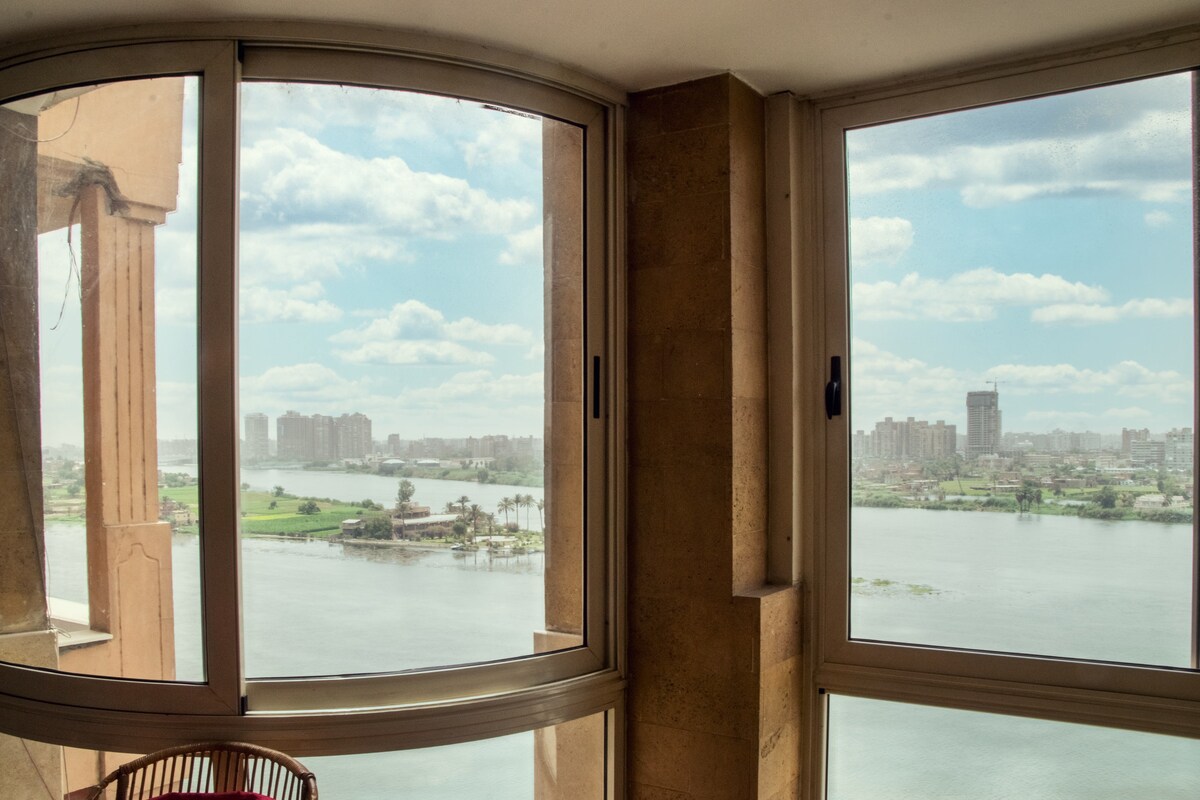 Large floor-to-ceiling windows provide expansive views of the Nile, showcasing the water and greenery beyond. The natural light illuminates the interior space, creating a bright and airy atmosphere within the room.