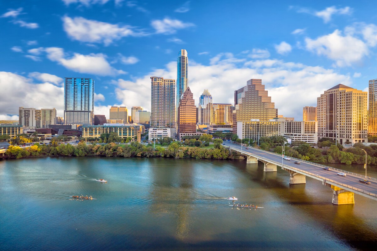 A city skyline is viewed from a waterfront perspective, showcasing modern high-rise buildings against a clear blue sky. Reflections of the structures shimmer on the water's surface, while a bridge spans across the river, with small boats visible in the foreground.