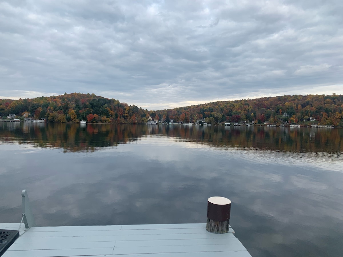 A calm lake reflects the colorful autumn foliage on the opposite shore, with gentle ripples across the water's surface. Overcast skies add a serene ambiance to the scene. The view is framed by a wooden dock in the foreground, featuring a cylindrical post.