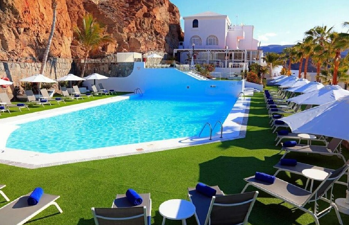 A tranquil pool area is surrounded by comfortable lounge chairs, with towels neatly arranged. Umbrellas provide shade, while lush green grass enhances the setting. Various plants and palm trees line the perimeter, and a visible rocky landscape adds natural beauty to the scene.