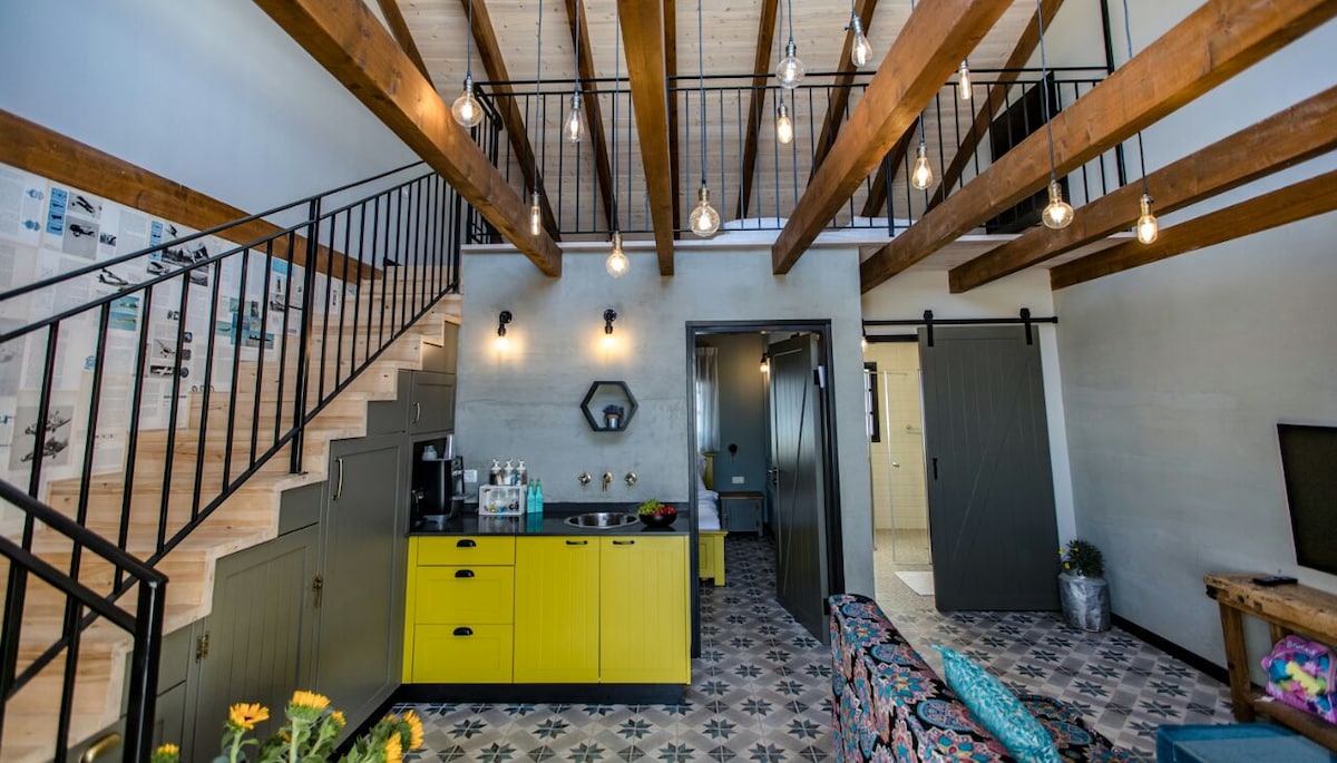 The open living area features a blend of industrial and rustic design elements. A vibrant yellow kitchen counter is complemented by decorative tiles on the floor. Exposed wooden beams and stylish pendant lights enhance the spacious layout, while a seating area invites relaxation.